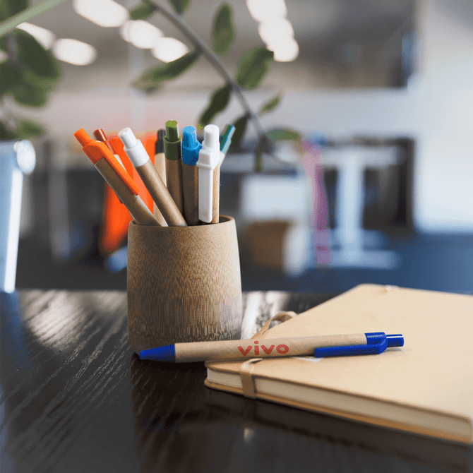 A wooden holder containing various coloured pens, including orange, green, and blue, beside a notebook on a desk.