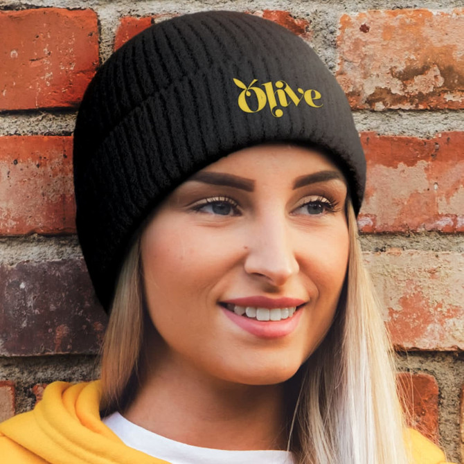 A black beanie with a ribbed texture, featuring a logo, worn by a smiling woman against a brick wall.
