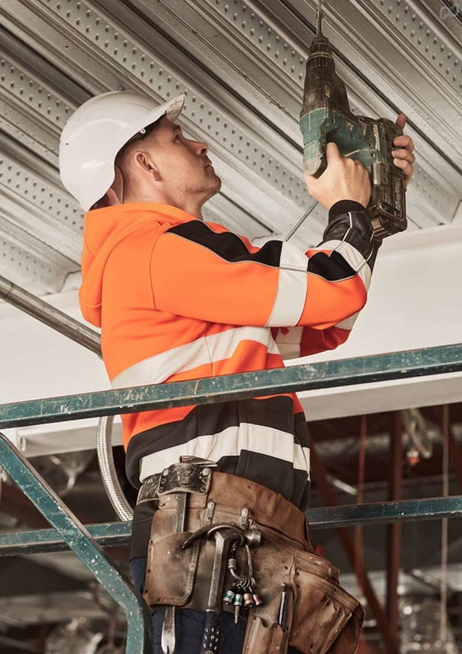 A unisex hi-vis full zip stretch taped hoodie in orange and black, worn by a worker using a drill on a scaffold.