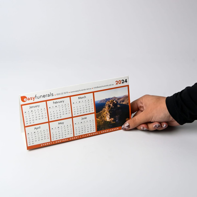 A desk calendar for 2024 featuring a scenic image, held by a hand, with orange and white colours.