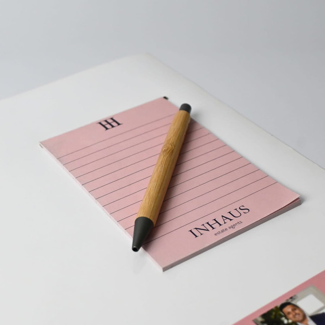 A pink notepad with lined pages and a wooden pen resting on top, featuring a logo.