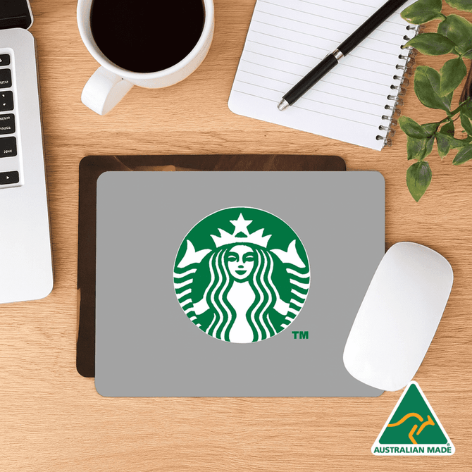 A grey mouse mat featuring a logo, placed on a wooden desk with a coffee cup, notebook, and plant.