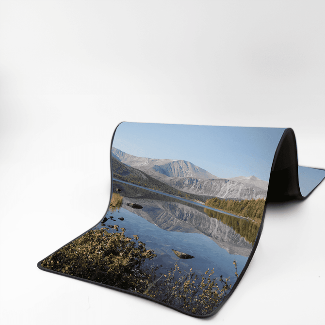 A rubber desk mat featuring a scenic landscape with mountains and a calm lake, predominantly in shades of blue and green.