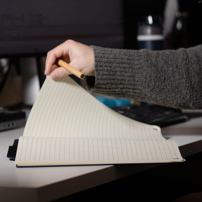 A hand holding a pencil opens a lined A4 notebook with a dark blue cover on a desk.