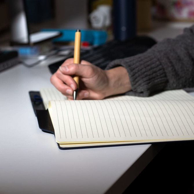 A hand holding a pen writes in a cream-coloured lined SunScribe notebook on a desk. The notebook has a blue cover.