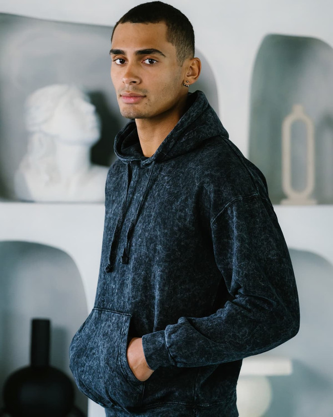 A model stands wearing a dark mineral wash pullover hoodie with a front pocket and a hood. The background features decorative items.