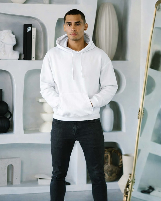 A man wearing a white hoodie and black jeans poses in a stylish indoor setting with modern decor.