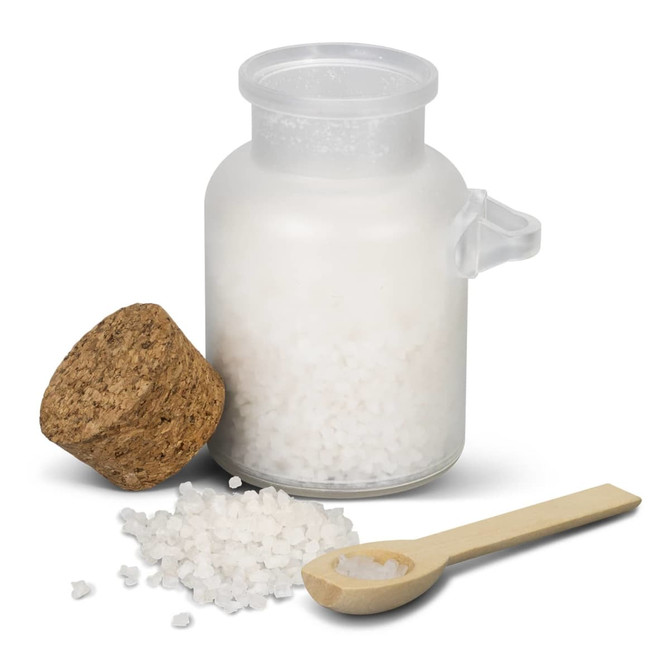 A clear glass jar filled with white bath salts, topped with a cork lid and accompanied by a wooden spoon.