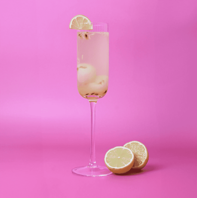A ribbed, clear glass champagne flute filled with a light beverage, garnished with a lemon slice, against a pink background.