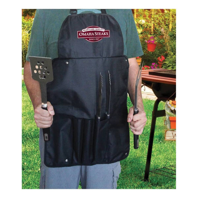 A black apron with pockets holds a spatula, tongs, and a barbecue fork, featuring a logo at the top.