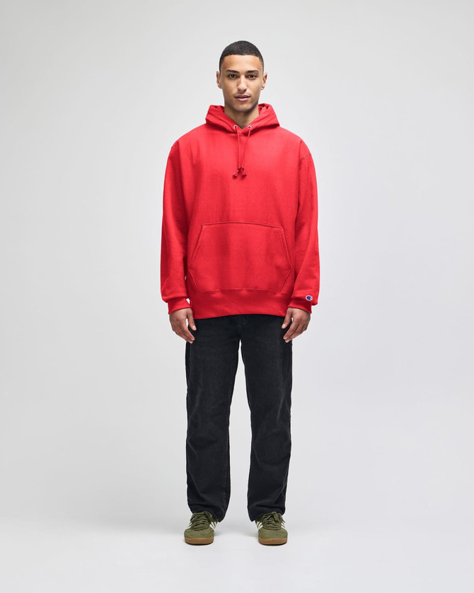 A red hooded sweatshirt with a front pocket and a logo, worn by a model standing against a neutral background.