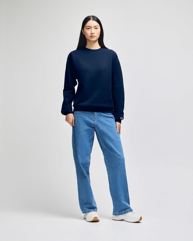 A model wearing a navy blue Champion Powerblend® adult crewneck sweatshirt paired with blue jeans and white shoes.