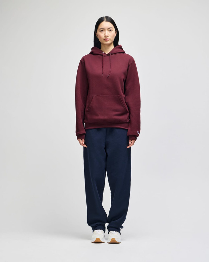 A maroon pullover hooded sweatshirt displayed on a model, paired with navy blue pants. The sweatshirt has a logo.