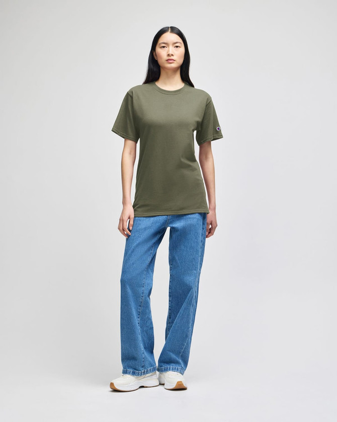 A model stands wearing a green Champion short-sleeve t-shirt paired with blue jeans and white sneakers.