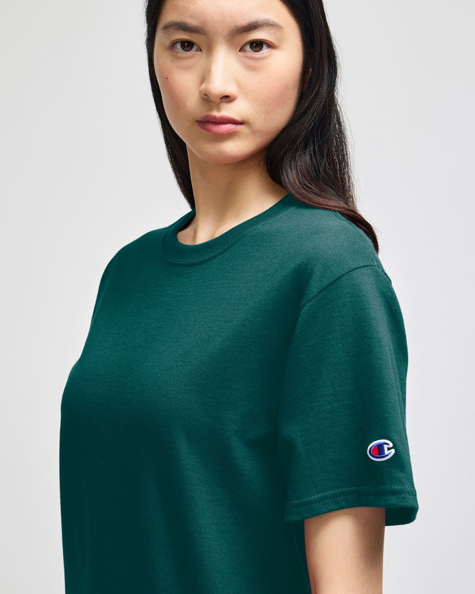 A model wearing a green Champion adult short-sleeve t-shirt, featuring a small logo on the sleeve.