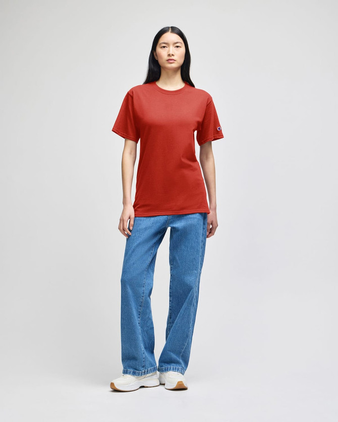A model wearing a red Champion adult short-sleeve t-shirt paired with blue jeans and white sneakers.