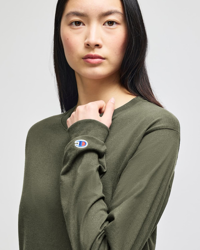 An olive green long-sleeve t-shirt featuring a small logo on the sleeve, modelled by a woman.