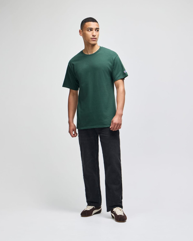 A green short sleeve t-shirt worn by a standing man, paired with dark pants and shoes. The shirt has a logo.