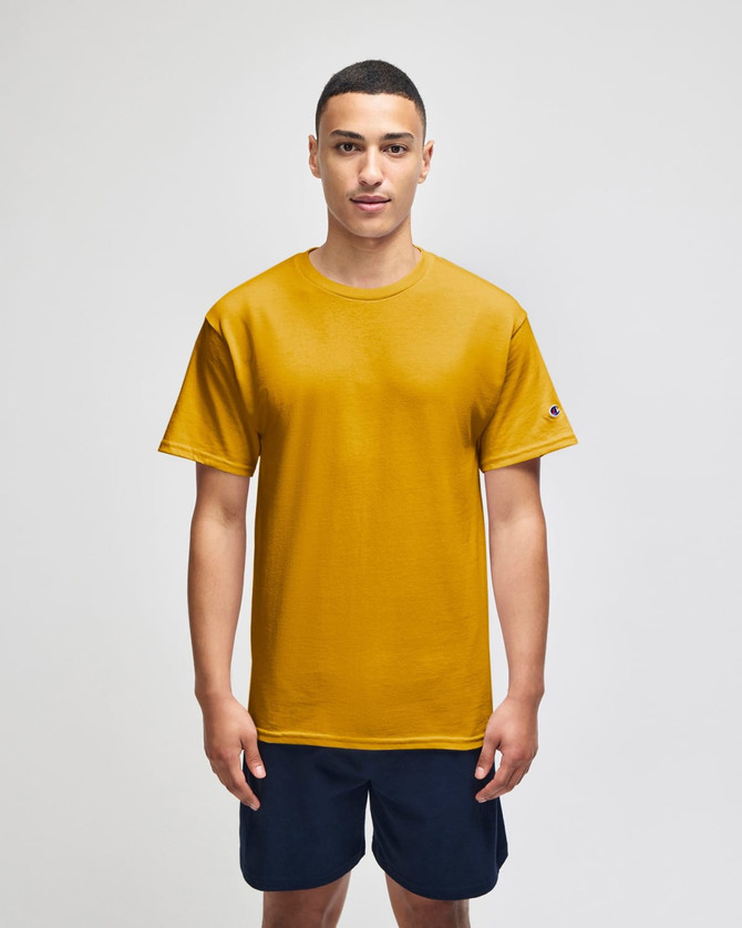 A man stands wearing a mustard yellow Champion Adult Jersey Short Sleeve T-Shirt with a logo, paired with navy shorts.