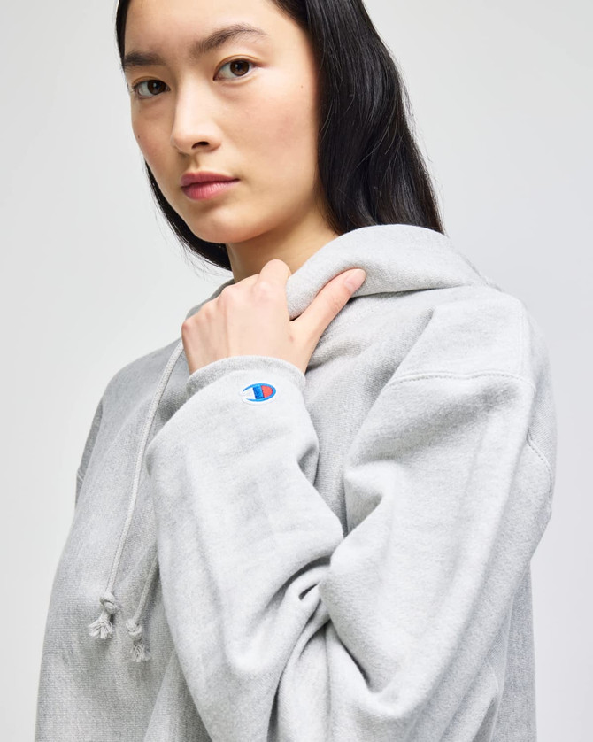 A grey cropped hoodie with a large hood and a logo on the sleeve, modelled by a woman.