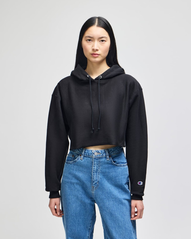 A black cropped hoodie with a hood and long sleeves, worn with blue jeans. It features a logo on one sleeve.