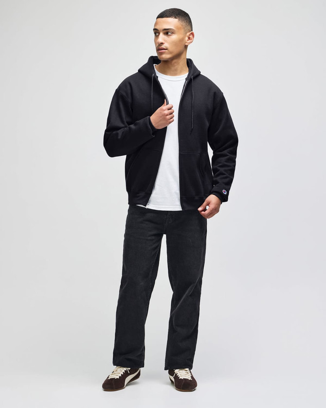 A model wearing a black full-zip hooded sweatshirt over a white t-shirt, paired with dark trousers and sneakers.