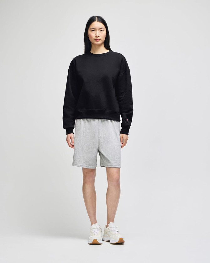A black crewneck sweatshirt worn with grey shorts, paired with white sneakers, featuring branding.