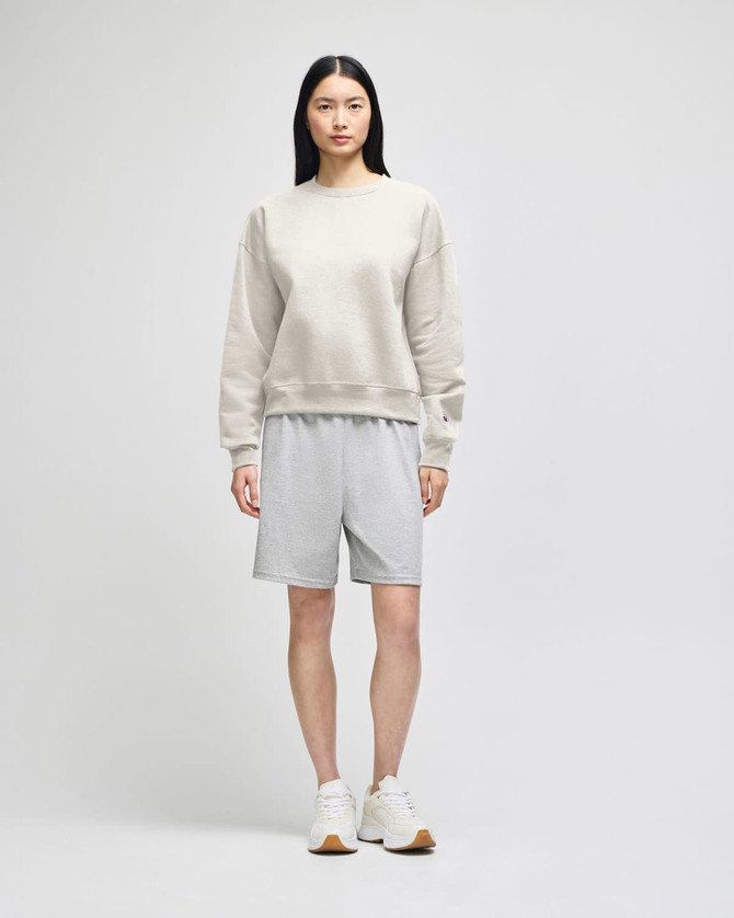 A model wearing a light grey crewneck sweatshirt and grey shorts, paired with white sneakers. The sweatshirt has a logo.