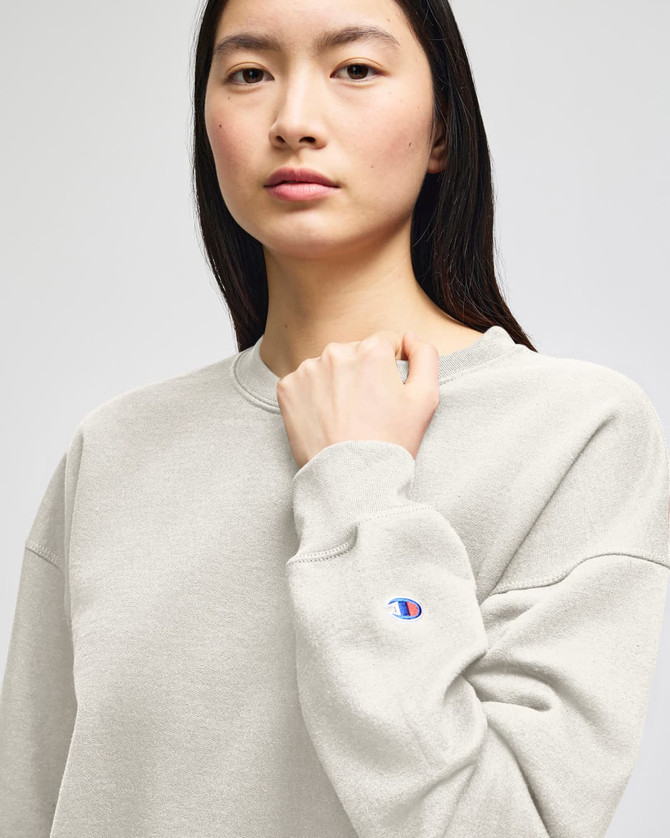A grey Champion Powerblend® women's crewneck sweatshirt with a logo on the sleeve, worn by a model.