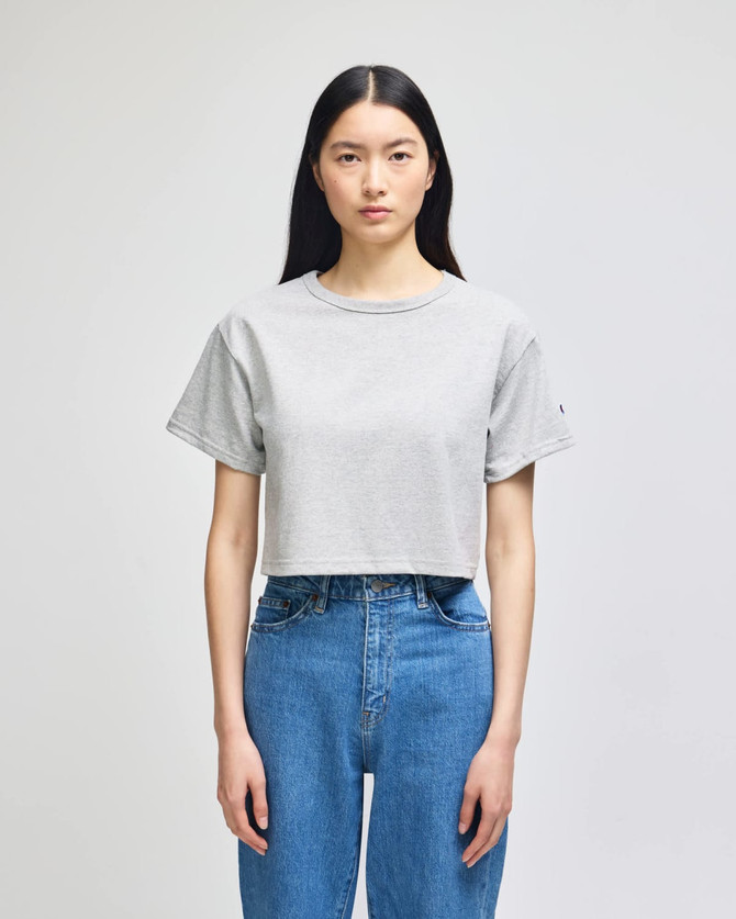 A cropped short sleeve jersey t-shirt in light grey, featuring a logo, paired with high-waisted blue jeans.