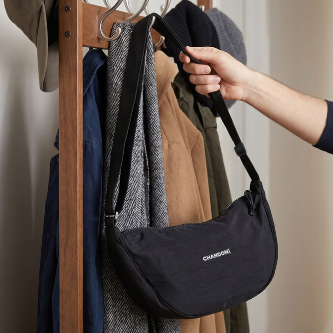 A black shoulder bag is being held on a coat rack, surrounded by jackets and hats. The bag has a logo.