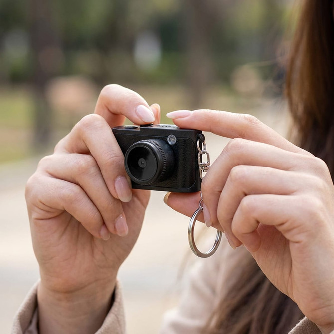 A compact black camera held in a person's hand, featuring a keychain attachment.