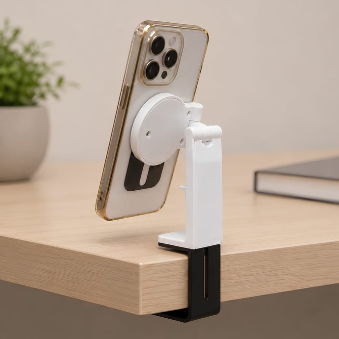 A white and black mobile phone stand holds a phone, attached to a wooden desk. A small plant and a book are in the background.