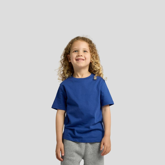 A child wearing a blue t-shirt, smiling and standing against a light grey background. The t-shirt features a logo.