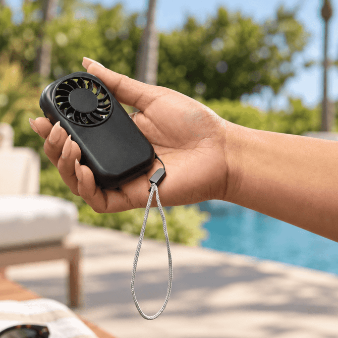 A handheld black mini pocket fan with a wrist strap, being held outdoors near a pool.