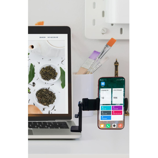 A mobile stand in black holding a smartphone beside a laptop, with a background of tea leaves and art supplies.