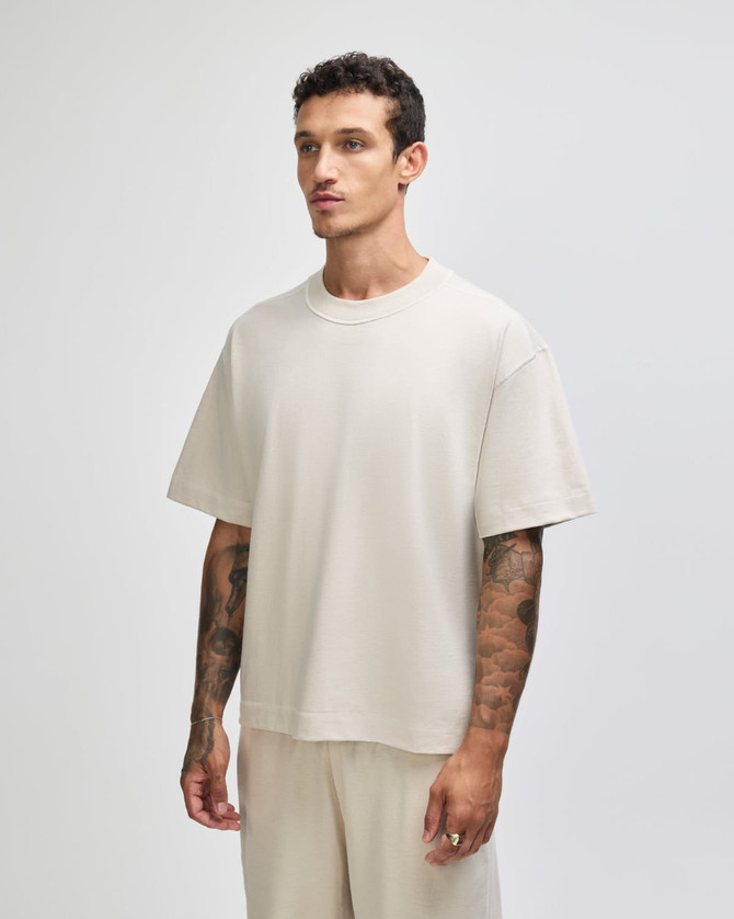 A boxy unisex t-shirt in a light beige colour, displayed on a model with tattoos. The shirt has a logo.