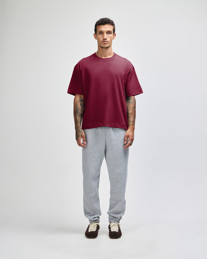 A model stands wearing a maroon heavyweight cotton unisex boxy t-shirt and grey sweatpants. The shirt has a logo.