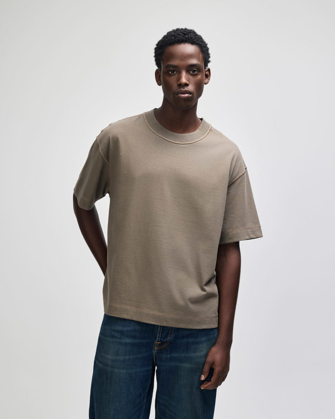A unisex boxy T-shirt in a muted olive green colour, worn by a model standing against a neutral background.