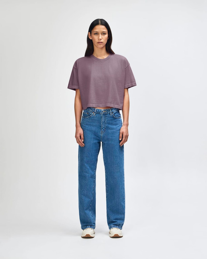 A heavyweight cotton women's crop boxy t-shirt in a muted purple, paired with blue jeans. The model stands against a plain background.