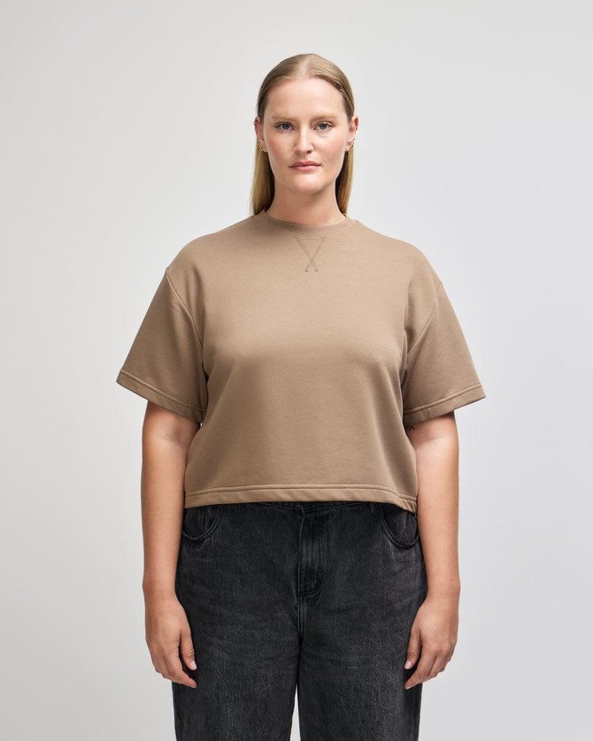 A mid-length boxy top in a tan colour, featuring short sleeves and a relaxed fit. The item has a logo.