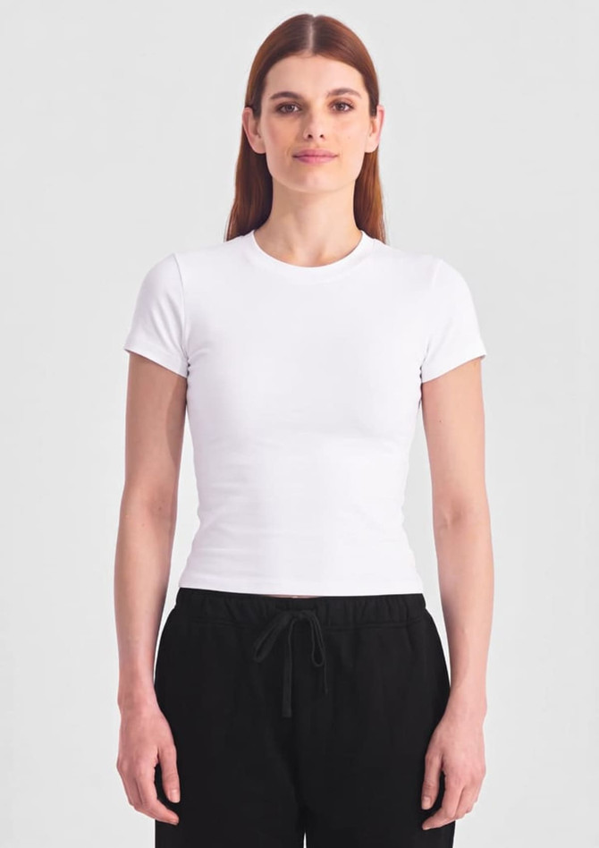 A white baby tee with short sleeves, paired with black trousers. The model stands against a neutral background.