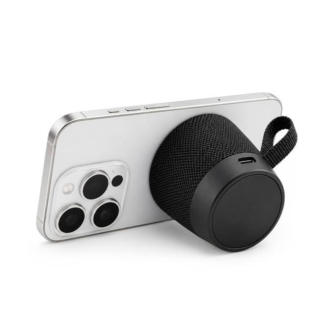 A black, cylindrical speaker with a textured surface, positioned against a smartphone. The speaker has a carrying strap.