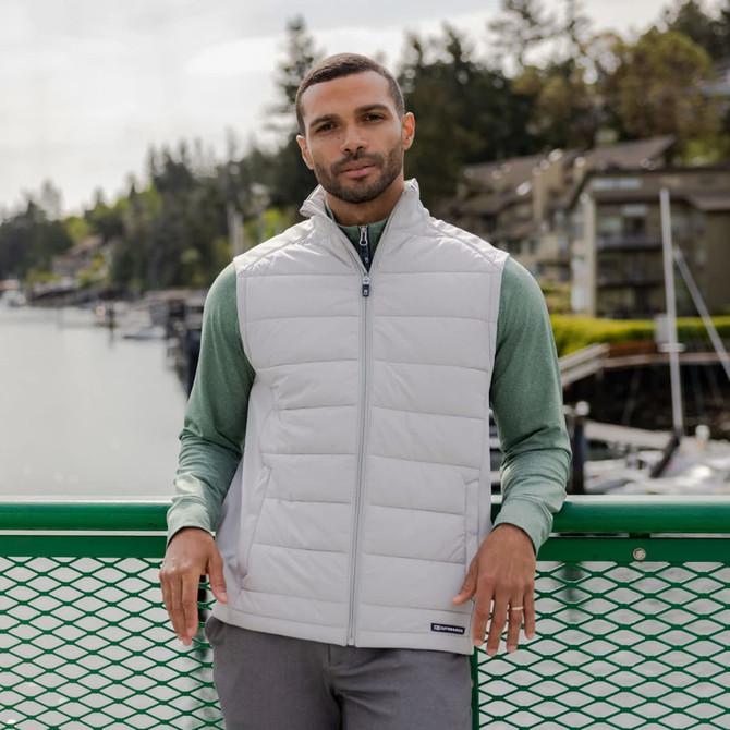 A men’s hybrid vest in light grey, featuring a zippered front and a logo, worn over a green long-sleeve top.