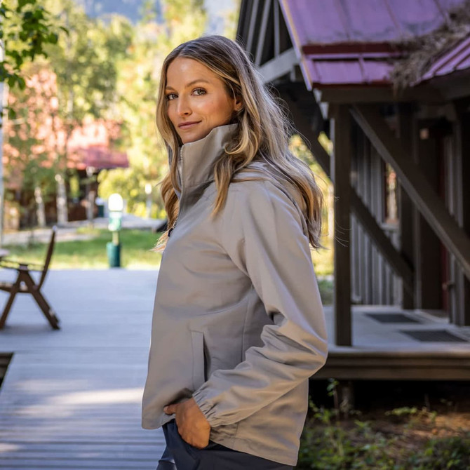 A light grey women's full zip jacket is worn by a model standing outdoors beside wooden buildings.