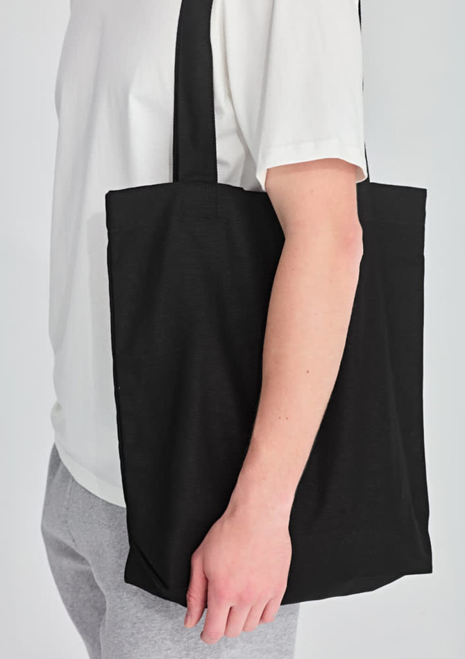A black canvas tote bag is held by a person wearing a white shirt and grey pants. The bag has a simple design.