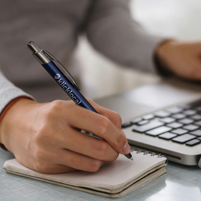 A metal pen in navy blue being used to write on a notepad next to a laptop. The pen features a branding logo.