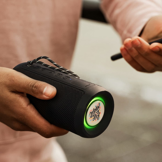 A Bluetooth speaker in black with a neon green light and a logo, held in a hand with a smartphone nearby.