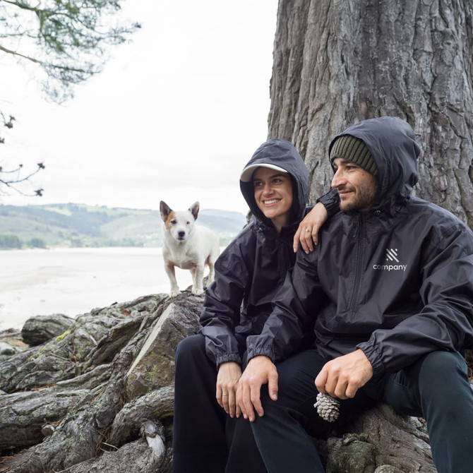 A black windbreaker worn by two people sitting by a tree, with a small dog nearby. The jackets have a logo on them.
