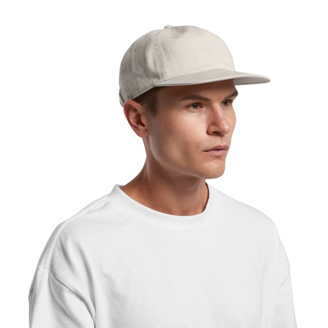 A light beige panel cap worn by a person, featuring a flat brim and a logo on the side.